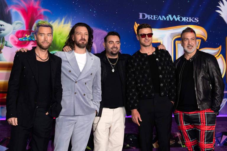 Cinco homens posam juntos em um evento, com um fundo colorido que inclui elementos gráficos da DreamWorks. Eles estão vestidos de maneira estilosa, com roupas que variam de ternos a camisetas e calças com padrões. 