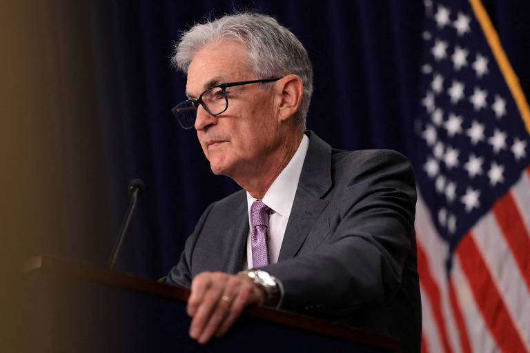 Foto de Jerome Powell, homem branco de cabelos grisalhos e óculos pretos, em uma tribuna. Veste terno e gravata violeta. Atrás dele, uma bandeira dos EUA.