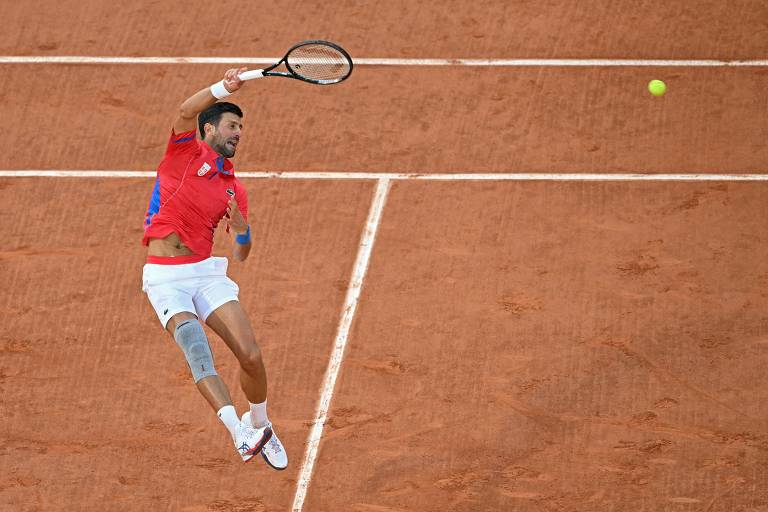 Um jogador de tênis está saltando para realizar um golpe com a raquete. Ele veste uma camiseta vermelha e calças brancas, com uma faixa na perna. A bola de tênis está visível no ar, enquanto o fundo é uma quadra de saibro.