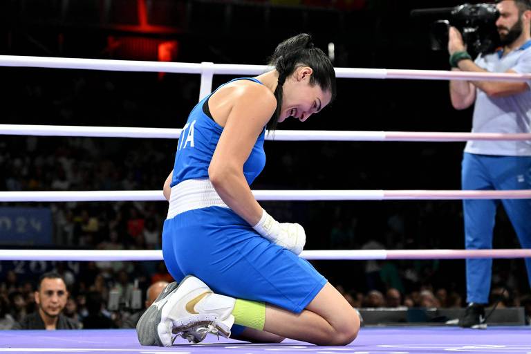 A imagem mostra uma boxeadora de joelhos no ringue, vestindo um uniforme azul com detalhes brancos. Ela parece estar em um momento de emoção, com a cabeça baixa e as mãos apoiadas nas pernas. Ao fundo, há um público visível e um cameraman registrando a cena.