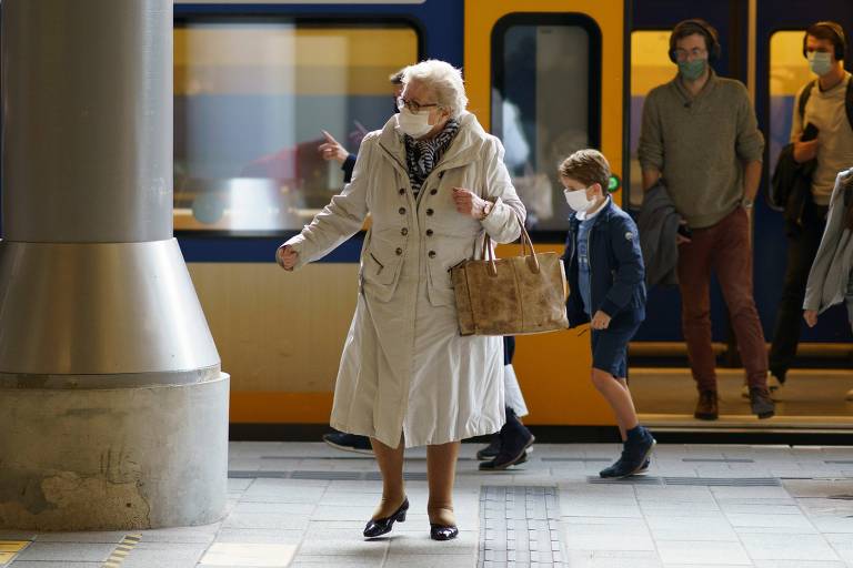 Mulher branca de cabelos brancos e curtos saindo de um trem. Ela está agasalhada com um sobretudo bege e carrega uma bolsa. Ela usa máscara facial descartável. O trem atrás é da cor amarela