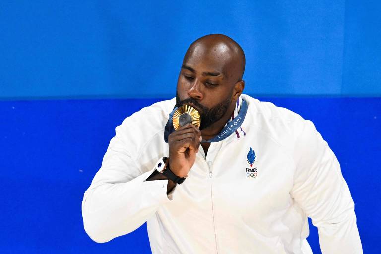 Um atleta está em pé, usando um casaco branco com o emblema da equipe olímpica da França. Ele está segurando uma medalha de ouro perto de seus lábios, como se estivesse prestando homenagem a ela. O fundo é azul, destacando a figura do atleta.
