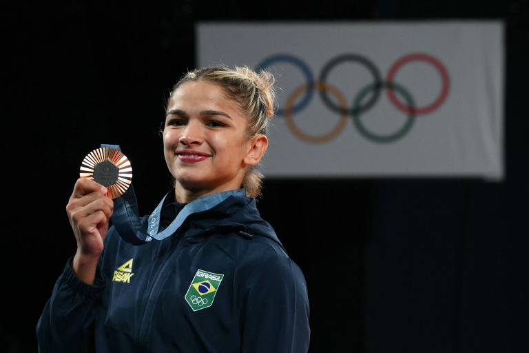 Uma atleta brasileira sorri enquanto segura uma medalha de bronze. Ela está vestindo um agasalho azul com o emblema do Brasil. Ao fundo, há os anéis olímpicos em cores diferentes, representando os Jogos Olímpicos.
