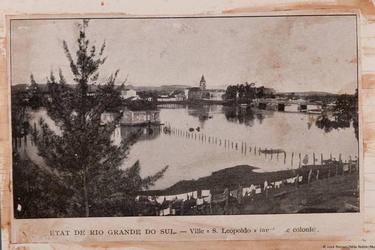 A imagem em preto e branco mostra uma vista panorâmica da cidade de São Leopoldo, no estado do Rio Grande do Sul. O cenário inclui um rio com casas à beira, uma ponte ao fundo e árvores em primeiro plano. Há também uma linha de postes de madeira no rio e roupas estendidas para secar na margem.