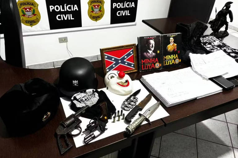 Na foto, armas, cadernos, dois livros Mein Kampf do ditador nazista Adolf Hitler e uma bandeira dos confederados norte-americanos está sob uma mesa em uma delegacia, como objetos recolhidos. Ao fundo, o símbolo da Polícia Civil.