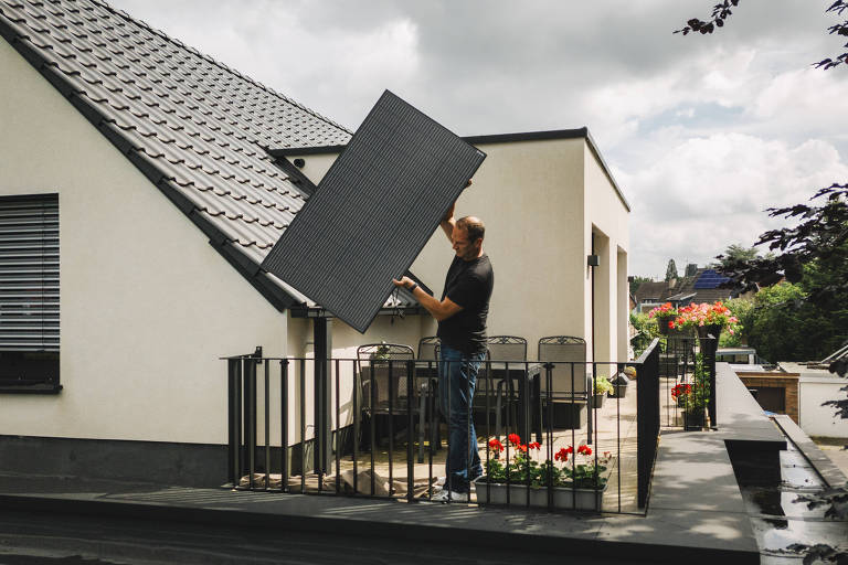 Um homem aparece na varanda de uma casa segurando um painel de energia solar. A casa é branca e tem telhado preto. 