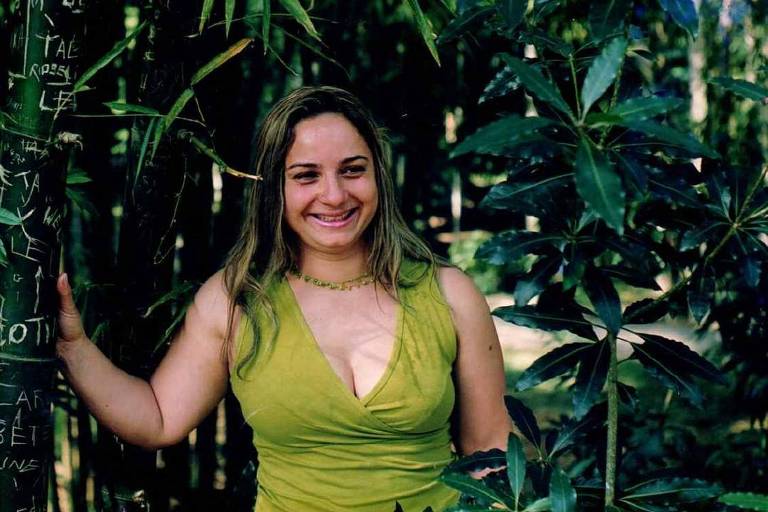 Uma mulher sorridente está posando ao lado de um tronco de bambu em uma área com vegetação densa. Ela usa uma blusa verde e tem cabelos longos e soltos. Ao fundo, há várias plantas verdes, incluindo folhas grandes e bambus.