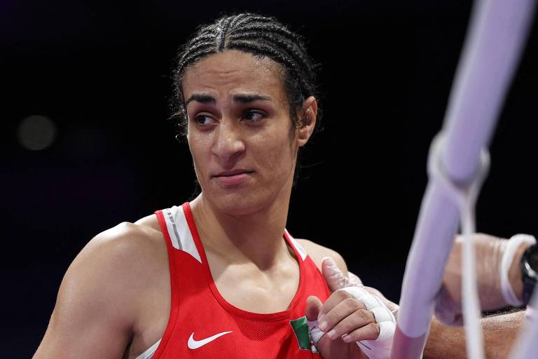 Imagem de uma atleta de boxe com cabelo trançado, vestindo um uniforme vermelho. Ela parece estar em um momento de reflexão ou concentração, com uma expressão facial séria. O fundo é desfocado, sugerindo um ambiente de competição.
