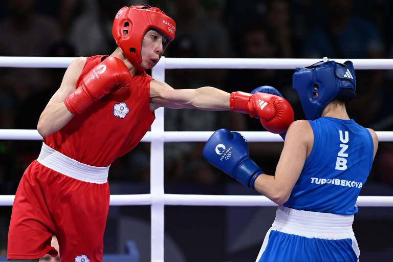 A imagem mostra uma luta de boxe entre dois atletas. Um lutador está vestido com um uniforme vermelho e um capacete vermelho, enquanto o outro está usando um uniforme azul e um capacete azul. O lutador vermelho está desferindo um soco em direção ao lutador azul, que está em posição defensiva. O cenário é uma arena de boxe, com um ringue e espectadores ao fundo.
