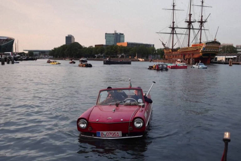 Fotografia mostra um carro vermelho navega em um canal. É fim de tarde e barcos são vistos ao fundo da imagem. 