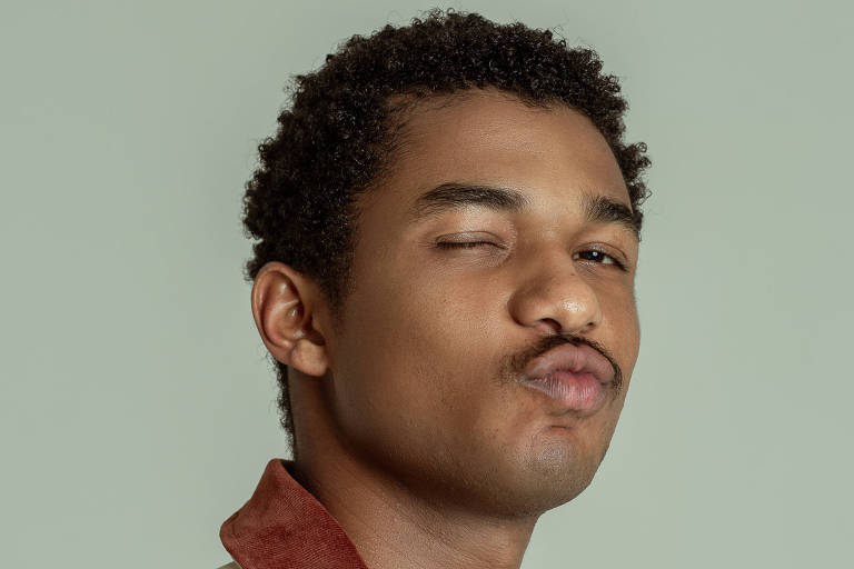 Um jovem está posando para a foto, com um leve sorriso. Ele tem cabelo cacheado e usa uma jaqueta de cor clara com detalhes em vermelho e estampas de folhas. O fundo da imagem é de uma cor verde suave.