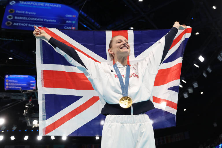 Uma atleta em um uniforme, segurando uma bandeira do Reino Unido acima da cabeça. Ela está sorrindo e exibindo uma medalha de ouro pendurada em seu pescoço. O fundo mostra uma arena com luzes e telas, sugerindo um evento esportivo.