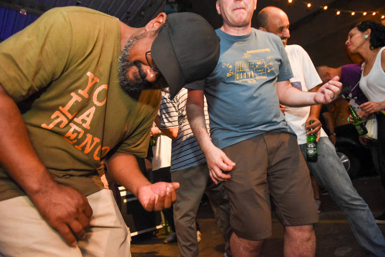 A imagem mostra um grupo de pessoas dançando em um ambiente festivo. Um homem com uma camiseta laranja escura e um boné está se movendo de forma animada, enquanto outro homem ao seu lado, vestindo uma camiseta azul e shorts, também está dançando. Ao fundo, há mais pessoas, algumas segurando bebidas. A iluminação é suave e o ambiente parece descontraído.