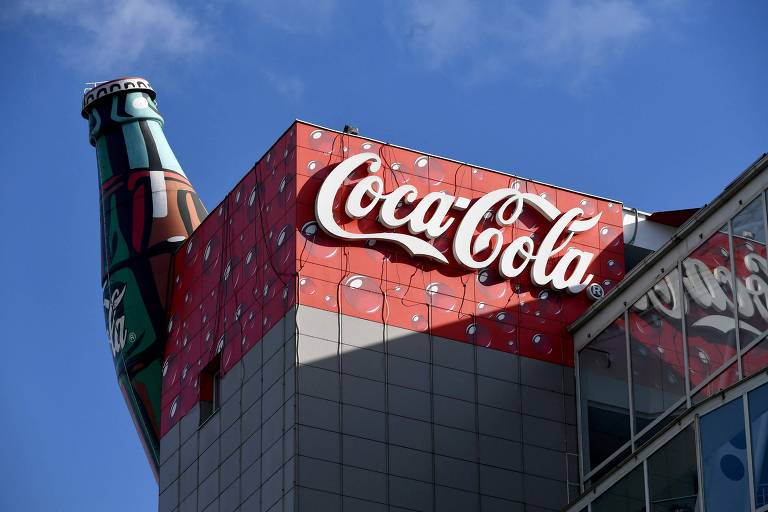 Fotografia mostra o alto de um prédio cinza com o topo vermelho e o símbolo da Coca-Cola parcialmente iluminado pela luz do sol. Parte de uma garrafa de refrigerante gigante é vista na parte de trás do edifício. No fundo, é possível ver o céu azul.
