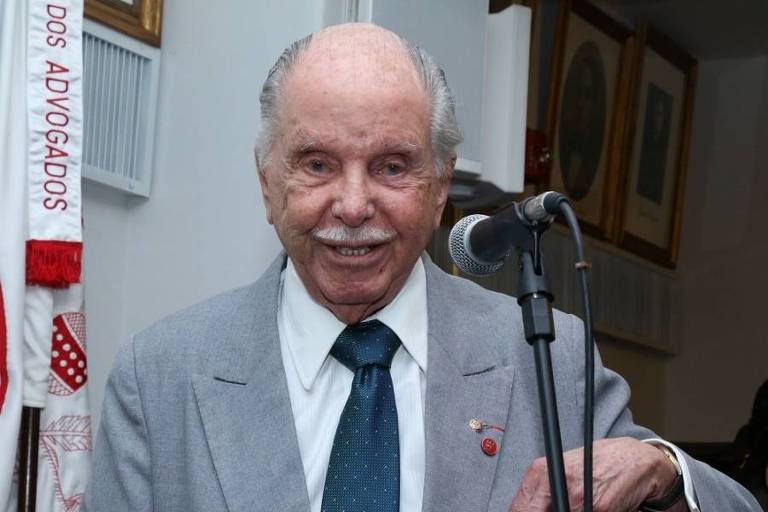 Um homem idoso, com cabelo grisalho e bigode, está sorrindo enquanto se apresenta em um evento. Ele usa um terno claro e uma gravata azul. Há um microfone à sua frente e ao fundo, é possível ver algumas molduras de quadros na parede.