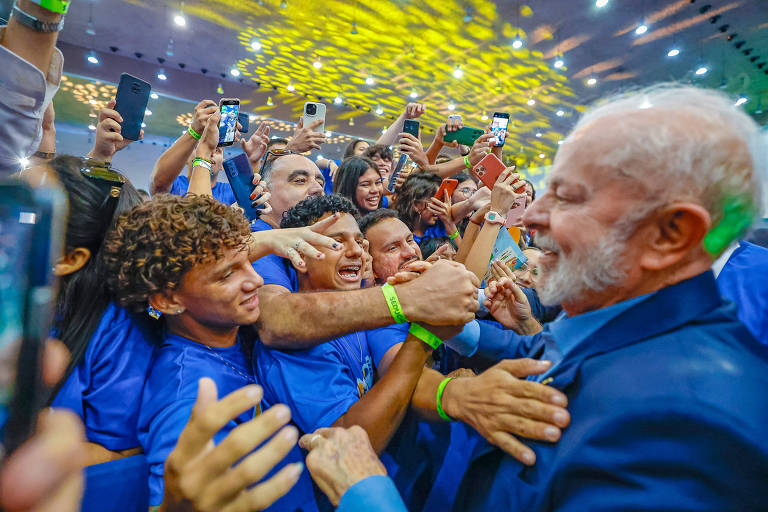 A imagem mostra um grupo de estudantes em um evento político, cercando o presidente Lula, que usa um terno azul. As pessoas estão vestidas com camisetas azuis e parecem animadas, algumas segurando celulares para registrar o momento. O ambiente é iluminado com luzes coloridas, criando uma atmosfera festiva.