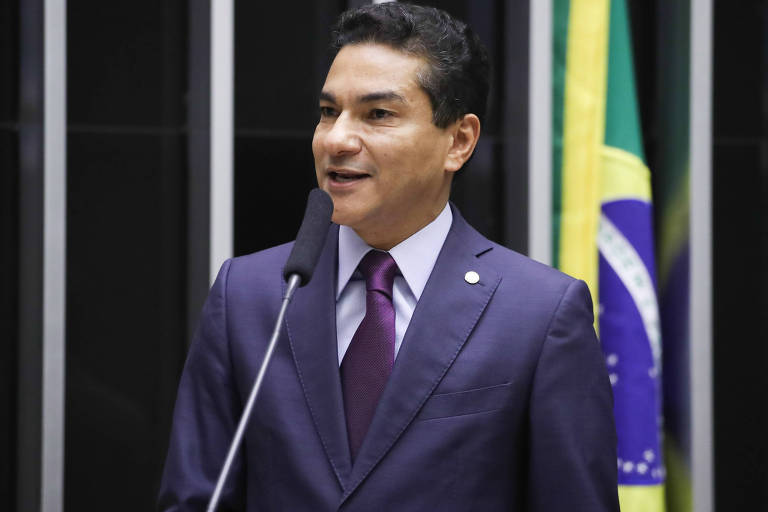 Um homem em um terno escuro e gravata roxa está falando em um microfone. Ele tem cabelo curto e liso, e está sorrindo enquanto se dirige a uma audiência. Ao fundo, há uma bandeira do Brasil visível, com as cores verde, amarelo e azul.