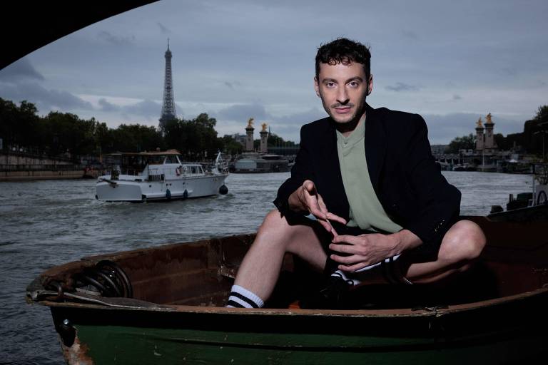 Um homem está sentado em um barco de madeira no rio Sena, com o corpo parcialmente exposto. Ele usa uma camiseta verde e um blazer preto, além de meias listradas. Ao fundo, é possível ver a Torre Eiffel e outros barcos navegando no rio, sob um céu nublado.