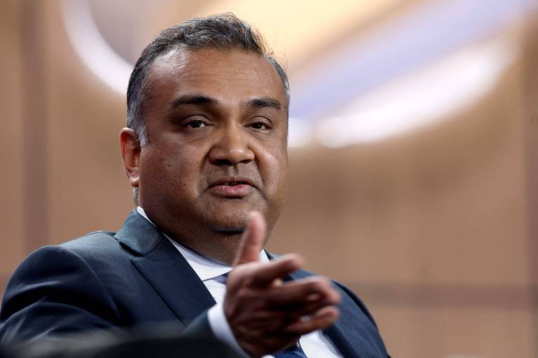 Um homem com cabelo curto e escuro, usando um terno escuro e uma camisa clara, está gesticulando com a mão direita enquanto fala. O fundo é desfocado, sugerindo um ambiente de conferência ou apresentação.