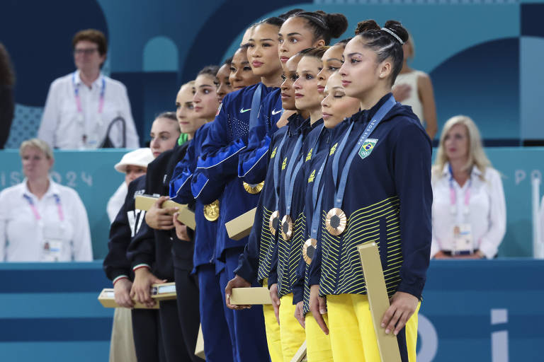 Um grupo de atletas de ginástica artística está em pé em um pódio, vestindo uniformes azuis e amarelos. Todas as atletas estão com medalhas ao redor do pescoço e segurando bastões. O ambiente é de competição, com pessoas ao fundo observando