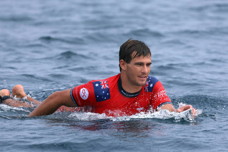 Foto mostra o surfista australiano Jack Robinson deitado sobre uma prancha dentro da água. Robinson é um homem branco com barba por fazer e cabelos loiros curtos. Ele veste uma camisa vermelha com o desenho da bandeira do seu país nos ombros.