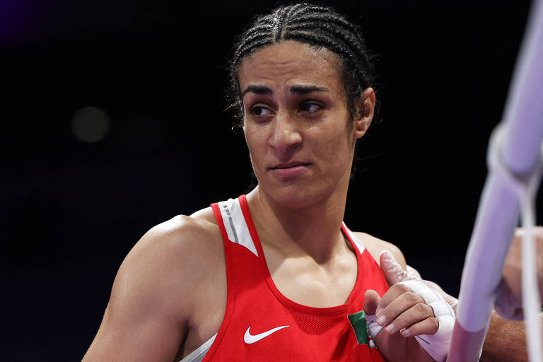 A imagem mostra uma boxeadora com cabelo trançado, vestindo um uniforme vermelho com detalhes brancos. Ela parece estar em um momento de pausa, com uma expressão de preocupação ou concentração. Sua mão direita está parcialmente visível, segurando uma fita ou um objeto, enquanto a mão esquerda é tocada por alguém fora do quadro. O fundo é desfocado, sugerindo um ambiente de competição.
