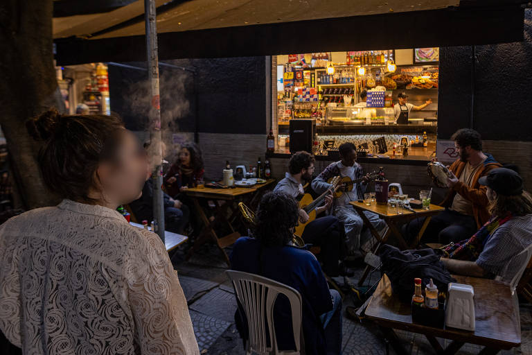 A imagem mostra um grupo de pessoas sentadas em mesas de madeira em um ambiente noturno, com uma mulher de costas observando e fumando. O grupo de músicos, composto por homens e mulheres, toca instrumentos, enquanto alguns clientes apreciam a música. A cena é animada e descontraída, com fumaça visível no ar