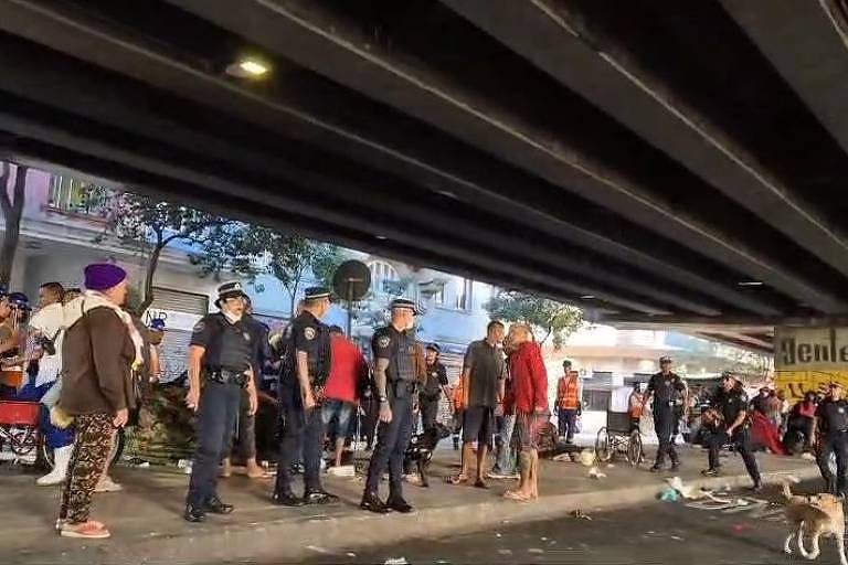 A imagem mostra uma cena sob um viaduto, onde há um grupo de guardas em uniformes azuis e várias pessoas, algumas sentadas e outras em pé. O ambiente parece desordenado, com lixo espalhado pelo chão. Há também bicicletas e um cachorro na cena