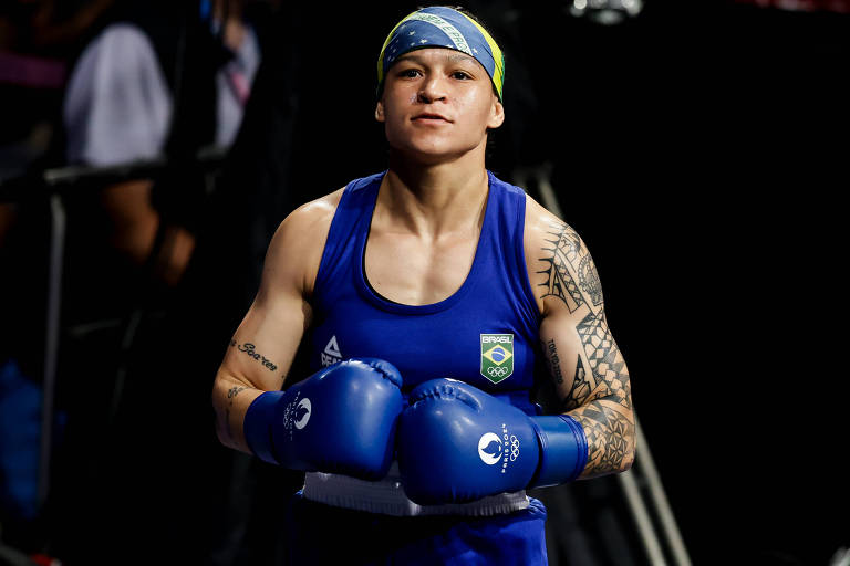 Foto mostra Bia Ferreira da cintura para cima. Ela esta com o uniforme de pugilista azul do Brasil, incluindo as luvas. Na cabeça, Bia, uma mulher com a pele clara, usa uma bandana com a bandeira brasileira. O fundo da imagem está escuro o que a deixa em evidência. 