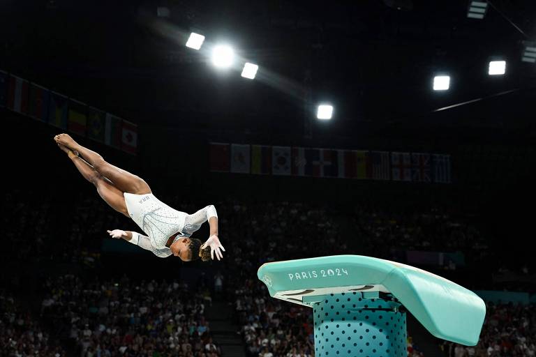 Uma ginasta está realizando um salto acrobático a partir de um trampolim. Ela está no ar, com o corpo em posição horizontal e os braços estendidos. O trampolim é de cor verde e está localizado em um ambiente de competição, com luzes brilhantes e uma plateia ao fundo.
