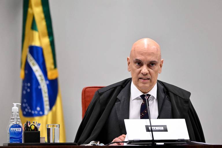 Imagem mostra um homem careca, de camisa social branca, gravata e uma capa preta nos ombros. Ele está sentado em uma cadeira vermelha e há um microfone na mesa à sua frente. Ao fundo, no lado esquerdo da imagem, há uma bandeira do Brasil.