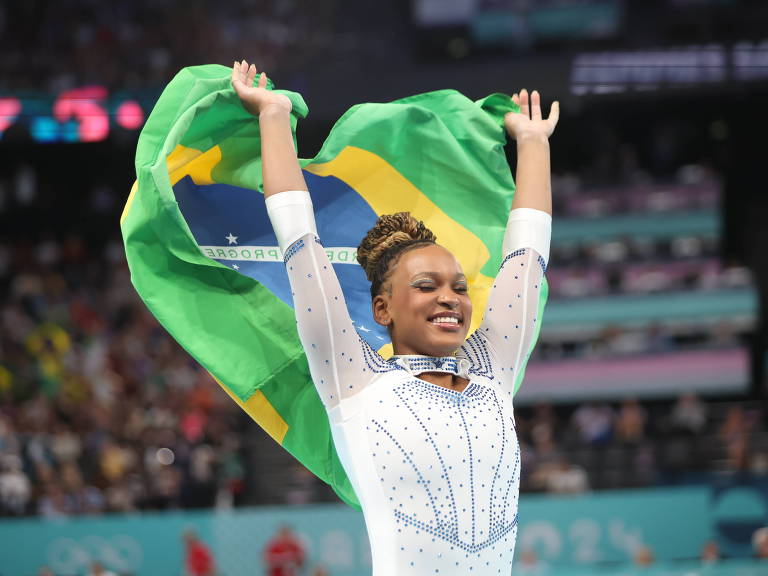 Vestindo collant branco, segurando a bandeira do Brasil e sorrindo, a ginasta brasileira Rebeca Andrade festeja a prata na prova do salto nas Olimpíadas de Paris-2024