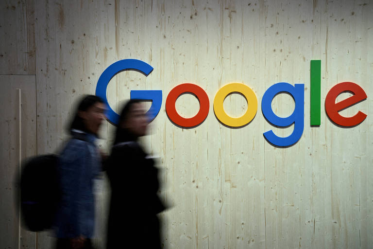 Vulto de duas pessoas desfocadas caminhando em frente a uma parede clara com o logo do Google. As pessoas usam roupas escuras e o logo do Google é colorido. 