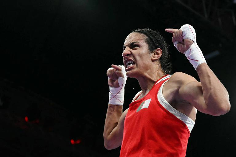 Um boxeador está em uma pose de celebração, com expressão intensa e determinada. Ele usa um uniforme de boxe vermelho e bandagens brancas nas mãos. O fundo é escuro, sugerindo um ambiente de competição, possivelmente um ringue de boxe.
