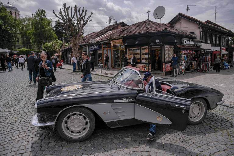 A imagem mostra um carro esportivo clássico, preto, estacionado em uma rua de paralelepípedos. O carro tem uma porta aberta e está em frente a uma loja com várias pessoas ao redor. Ao fundo, há edifícios com telhados de madeira e árvores. O céu está nublado.
