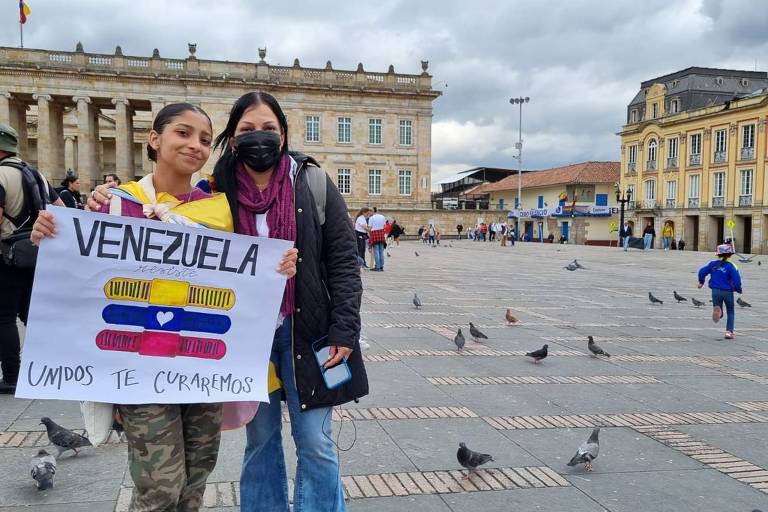 Duas mulheres posam em uma praça, segurando um cartaz que diz 'VENEZUELA' com as cores da bandeira venezuelana e a frase 'JUNTOS TE CURAREMOS'. Ao fundo, há um edifício histórico e várias pessoas e pombos na praça.