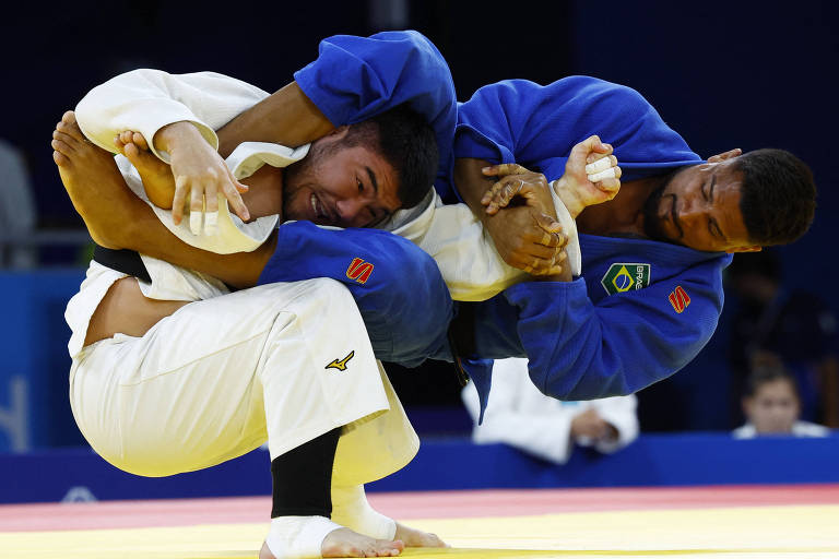 Foto mostra judoca brasileiro aplicando um golpe no adversário cazaquistanês. Léo, um homem negro com bigode e cabelos curtos pretos, usa kimono azul com bandeira do Brasil. Ele está suspenso no ar enquanto dá uma chave de braço no adversário, um homem asiático com barba e cabelo preto arrepiado, que veste um kimono branco. 