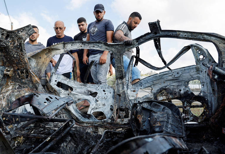 A imagem mostra um carro queimado, com a estrutura metálica exposta e danificada. Várias pessoas estão em pé ao redor do carro, observando os danos. O céu está claro ao fundo, e a cena parece ser ao ar livre