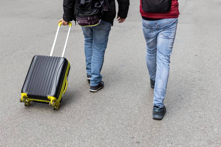 Duas pessoas caminham, de costas. Uma delas carrega uma mala de rodinhas preta