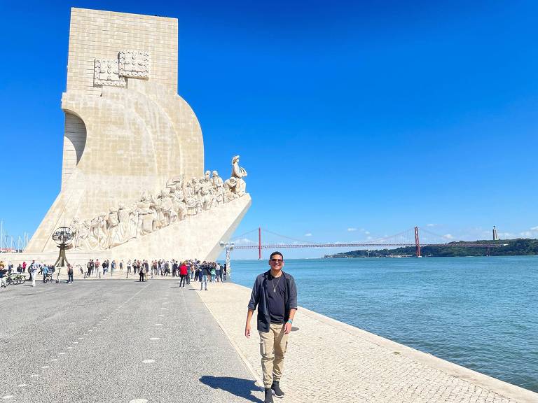 A imagem mostra uma pessoa caminhando em frente ao Monumento aos Descobrimentos, em Lisboa. O monumento é grande e apresenta figuras esculpidas que representam exploradores e navegadores. Ao fundo, há um rio e uma ponte visível. O céu está limpo e azul, e há várias pessoas ao redor do monumento