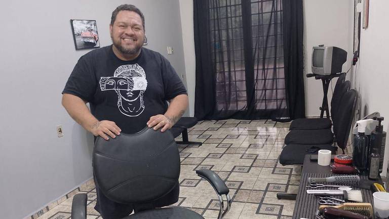 Um homem sorridente está em pé ao lado de uma cadeira de barbearia em um ambiente de barbearia. Ele usa uma camiseta preta com um desenho. O espaço é bem iluminado, com piso de azulejos e paredes em tons claros. Ao fundo, há uma janela com cortinas pretas e algumas cadeiras de espera. Em uma mesa próxima, há produtos de cabelo e ferramentas de barbearia