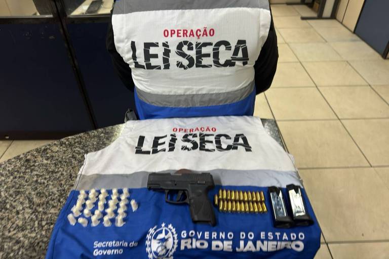 Sobre uma mesa, um colete azul e branco da Operação Lei Seca. Em cima do colete, organizados, estão pinos de cocaína, que são objetos cônicos brancos, uma pistola preta e munição