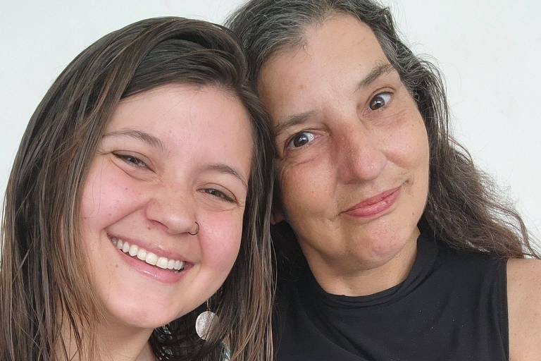 A imagem mostra duas mulheres posando juntas para uma selfie. Ambas estão sorrindo. A mulher à esquerda tem cabelo longo e castanho claro, enquanto a mulher à direita tem cabelo longo e castanho escuro, com algumas mechas grisalhas