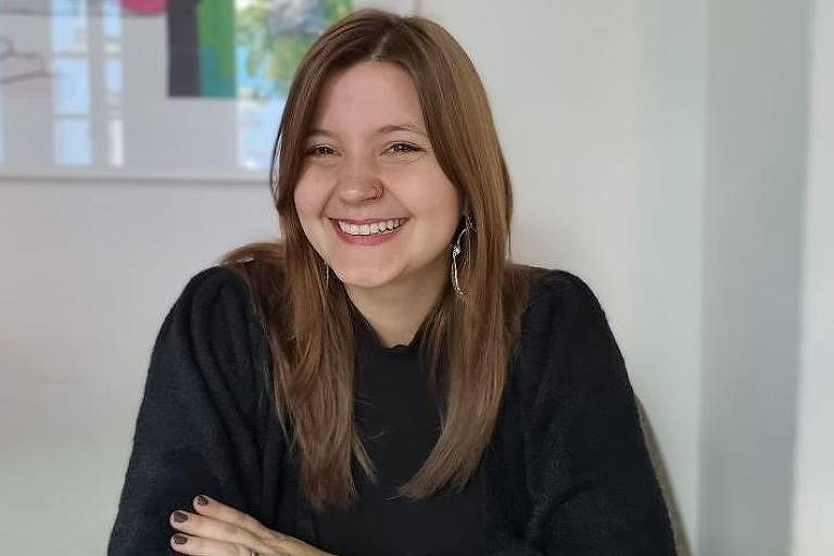 A imagem mostra uma mulher com cabelo castanho claro e liso, usando uma blusa preta. Ela está sorrindo e com os braços cruzados sobre a mesa