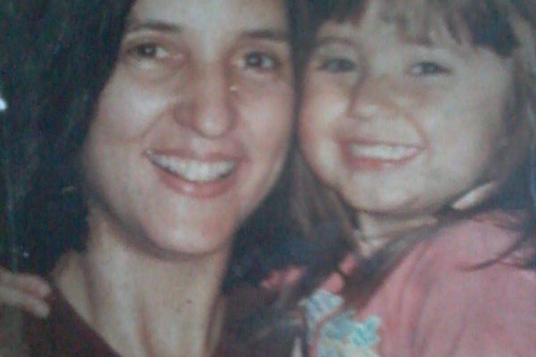 A imagem mostra uma mulher sorridente segurando uma menina pequena em seu colo. Ambas estão olhando para a câmera e sorrindo. A mulher tem cabelo escuro e liso, enquanto a menina tem cabelo castanho claro e ondulado. Elas estão em um ambiente com iluminação suave, e a expressão delas transmite felicidade e carinho