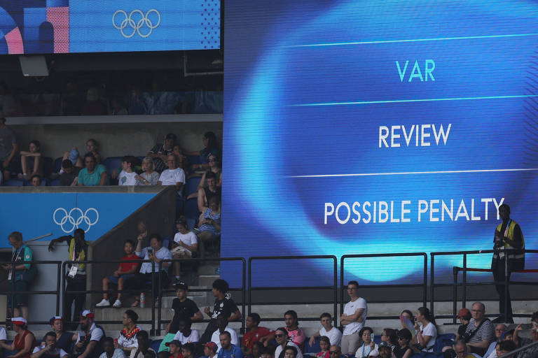 A imagem mostra uma tela grande em um evento esportivo, provavelmente durante os Jogos Olímpicos. Na tela, estão as palavras 'VAR', 'REVIEW' e 'POSSIBLE PENALTY', indicando que uma revisão de vídeo está em andamento para uma possível penalidade. Ao fundo, há uma plateia com várias pessoas assistindo ao evento.
