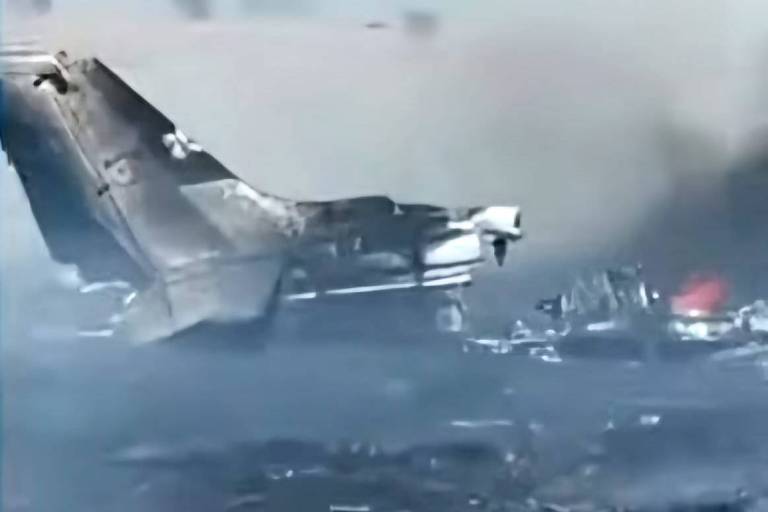 A imagem mostra os destroços de um avião em chamas, com fumaça densa ao fundo. A parte traseira da fuselagem está visivelmente danificada, e há uma área de fogo em meio aos destroços