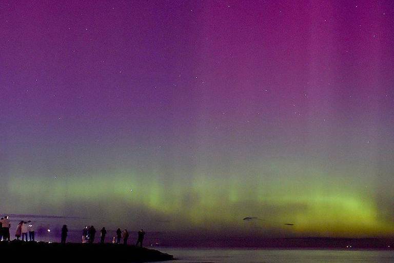 Foto colorida mostra céu estrelado com 'ondas de luz' verdes, roxas e azuis sobre o mar