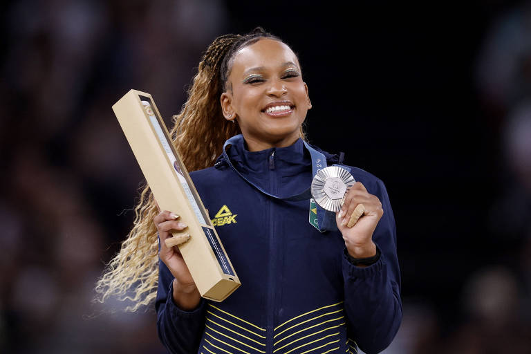 Uma atleta sorridente segura um troféu longo em uma mão e uma medalha prateada na outra. Ela está vestindo uma jaqueta esportiva azul com detalhes em amarelo e tem cabelo longo preso em um rabo de cavalo. O fundo é desfocado, sugerindo um ambiente de competição.