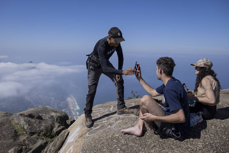 Três pessoas estão sentadas em uma rocha no topo de uma montanha, com um céu azul ao fundo. Um dos indivíduos, de pé, está passando um celular para outro que está sentado. A terceira pessoa, também sentada, observa. A paisagem ao fundo mostra nuvens abaixo da montanha.